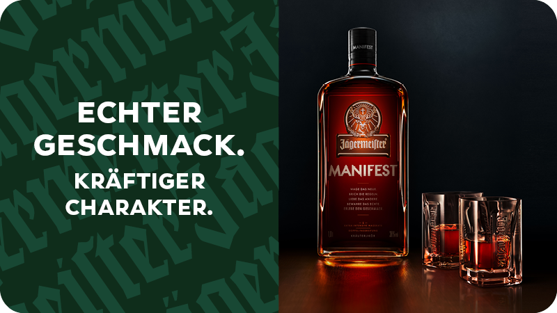 Im Vorgrund steht eine angerissene Flasche Jägermeister Manifest und im Hintergrund ist der untere Teil einer Person mit Jeans und Lederschuhen zu sehen